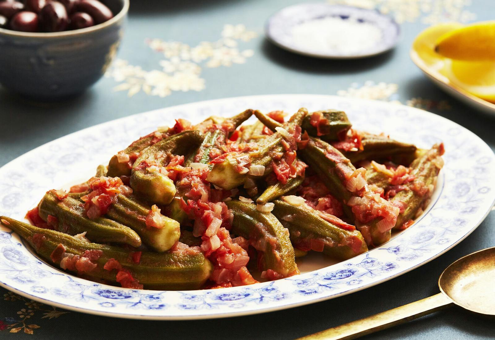 Okra with tomatoes, lemon wedges and olives atop navy tablecloth.