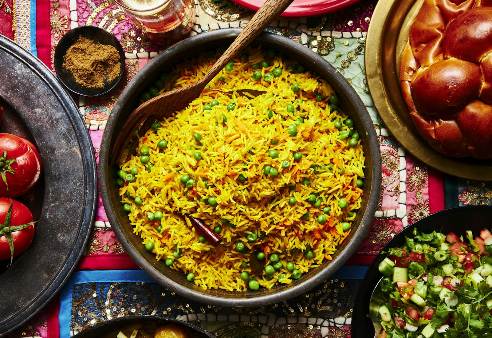 Pilau in large bowl with wooden serving spoon alongside challah, spice mix, beer, side salad and mahasha.