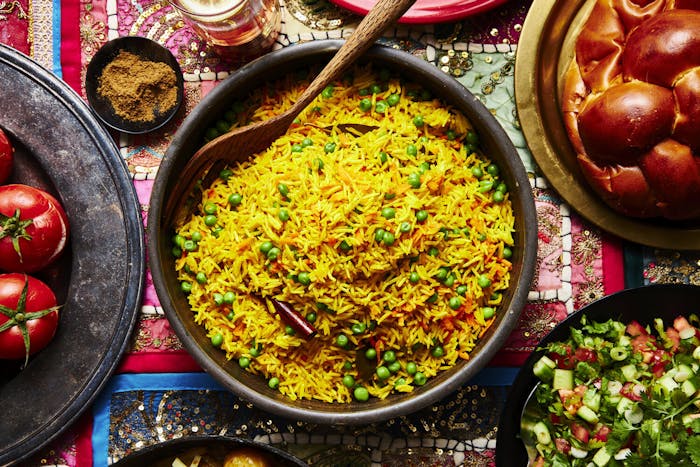 Pilau in large bowl with wooden serving spoon alongside challah, spice mix, beer, side salad and mahasha.