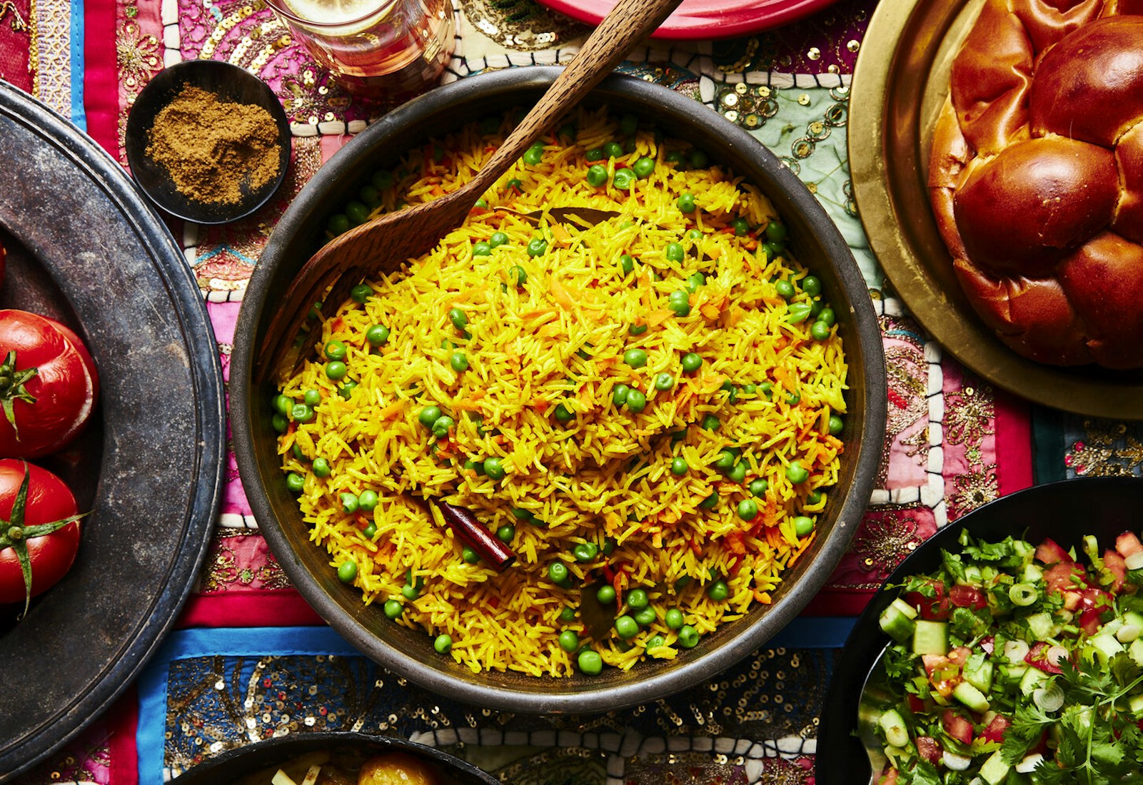 Pilau in large bowl with wooden serving spoon alongside challah, spice mix, beer, side salad and mahasha.