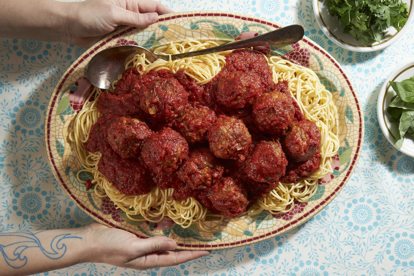 Meatballs with red sauce on bed of spaghetti, bowls with parsley and basil atop blue tablecloth.