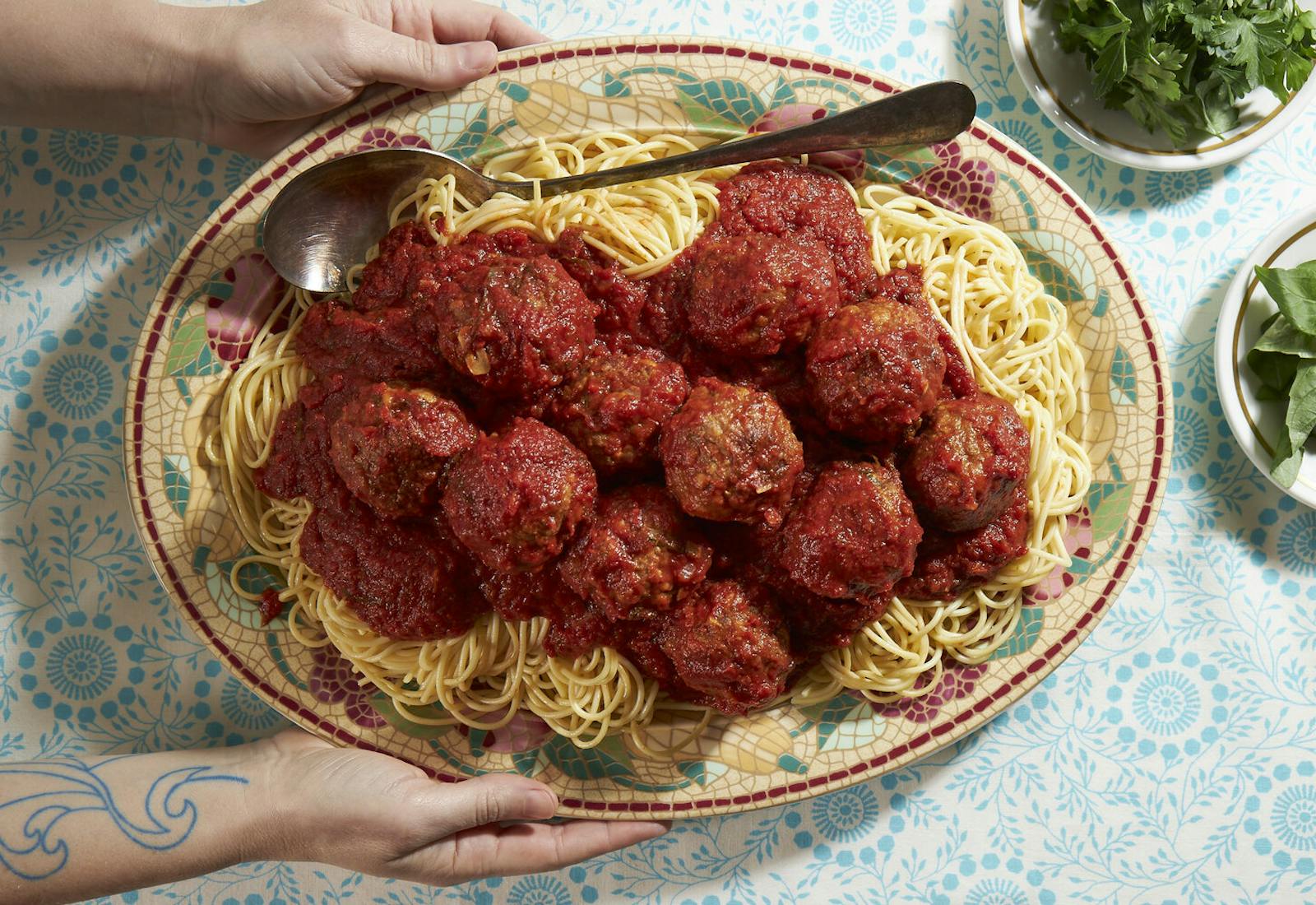 Meatballs with red sauce on bed of spaghetti, bowls with parsley and basil atop blue tablecloth.