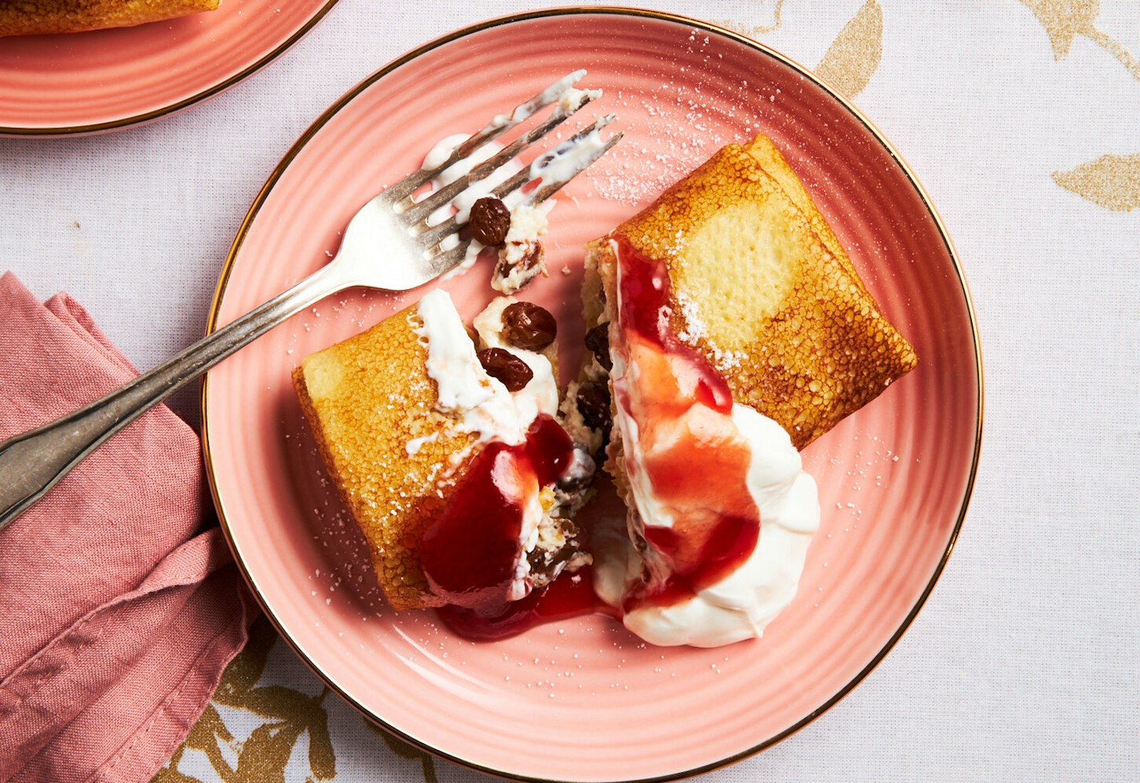 Nalesniki with jam and sour cream on salmon plate atop white and gold tablecloth.