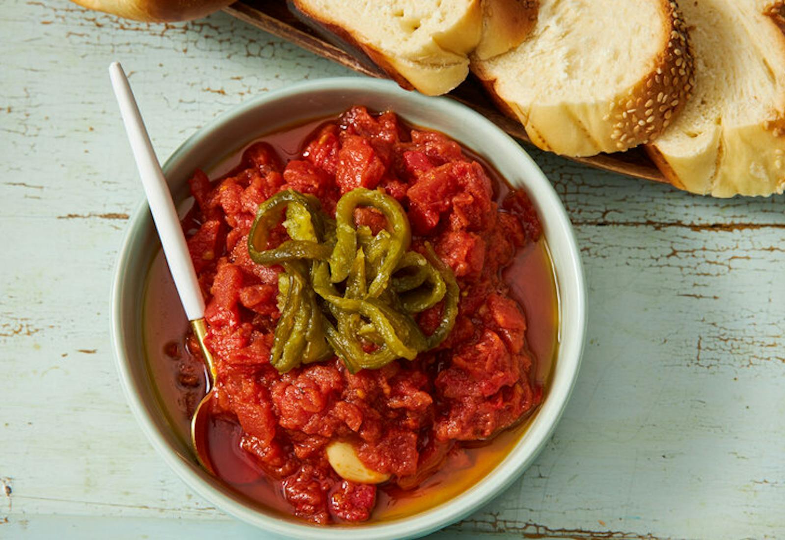 Bowl of matbucha garnished with roasted jalapeños, slices of challah atop blue wooden surface.