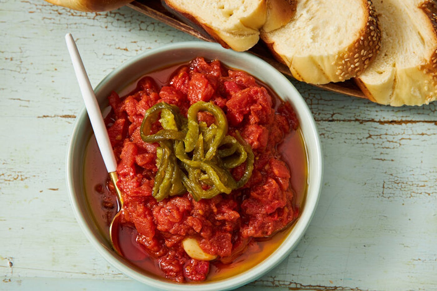 Bowl of matbucha garnished with roasted jalapeños, slices of challah atop blue wooden surface.