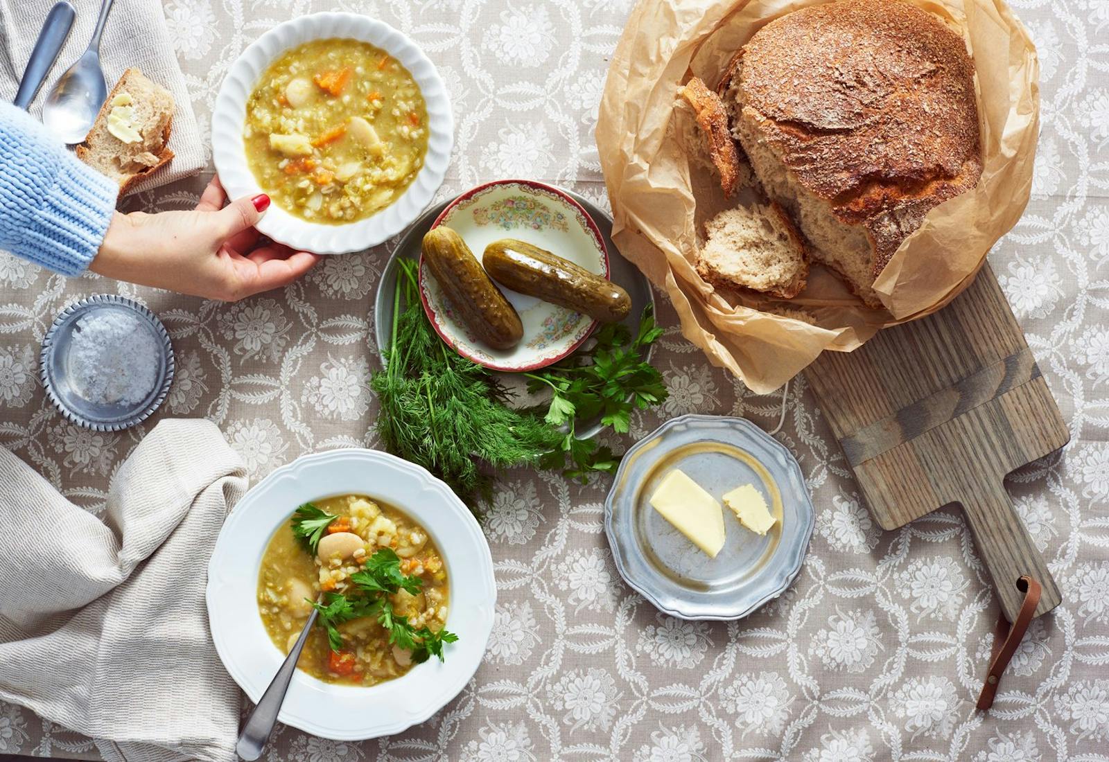 Hand placing bowl of soup onto table surrounded by fresh dill, pickles, butter and Lithuanian sour rye bread on wooden cutting board.