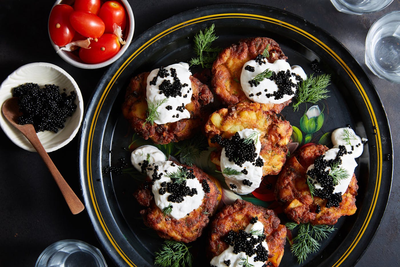 White fish and potato latkes with dollops of horseradish sour cream and black caviar, garnished with dill. Alongside bowls of cherry tomatoes and caviar atop black surface. 