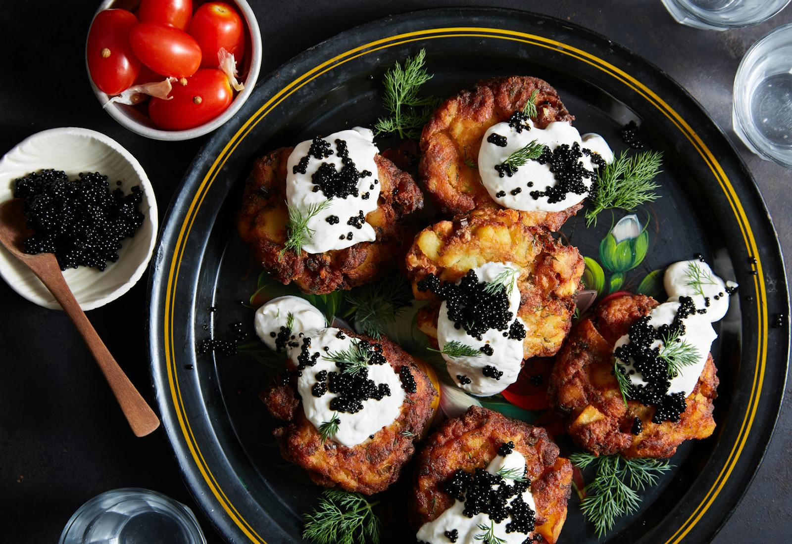 White fish and potato latkes with dollops of horseradish sour cream and black caviar, garnished with dill. Alongside bowls of cherry tomatoes and caviar atop black surface. 