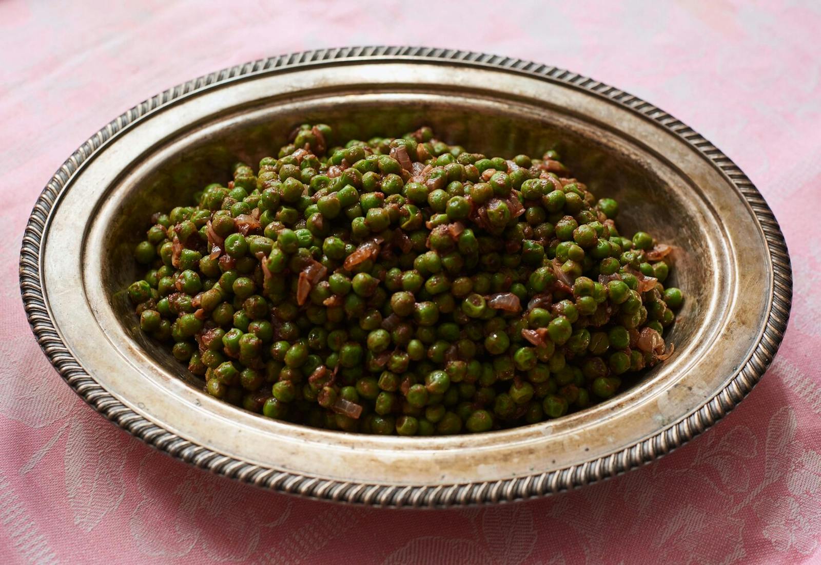 Peas with allspice in bronze serving bowl atop pink tablecloth.
