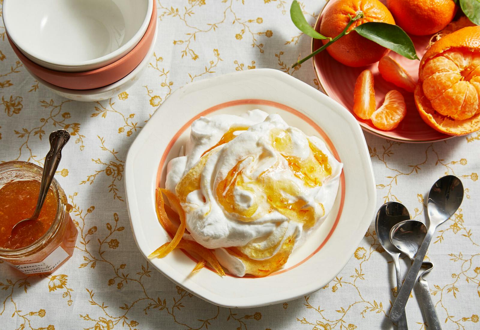 Meringue swirled with orange marmalade alongside bowl of fresh oranges, jar of marmalade and serving bowls, atop orange floral tablecloth.