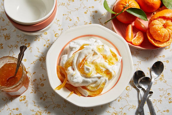 Meringue swirled with orange marmalade alongside bowl of fresh oranges, jar of marmalade and serving bowls, atop orange floral tablecloth.