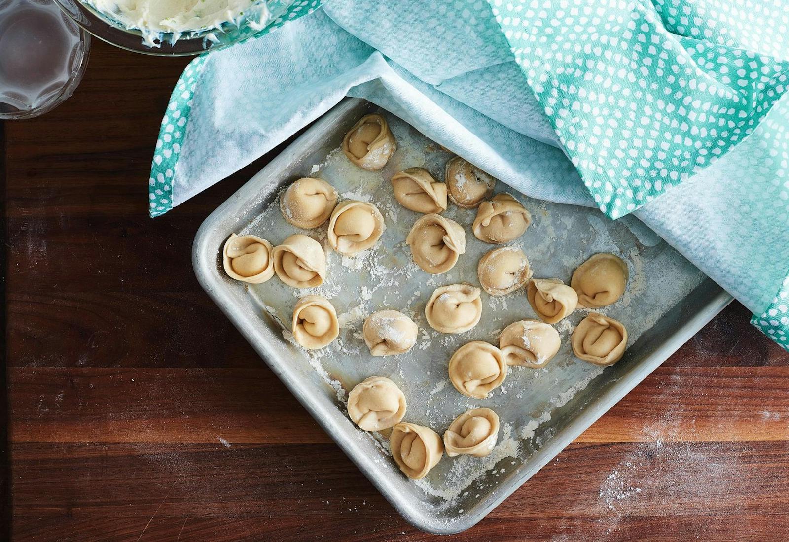 Flour-coated baking pan with uncooked cheese vareniki, blue and white kitchen cloth draped over dumplings, atop wooden surface.