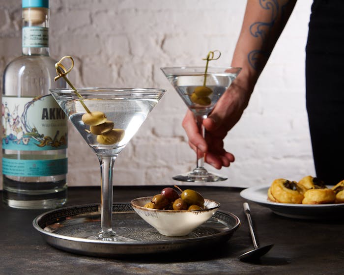 Chef serving dirty martinis alongside bowl of olives and bottle of Akko gin atop grey table