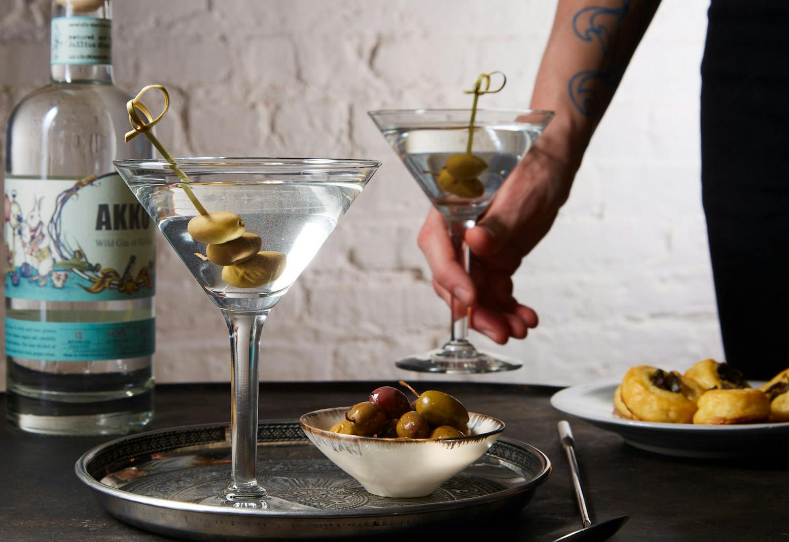 Chef serving dirty martinis alongside bowl of olives and bottle of Akko gin atop grey table