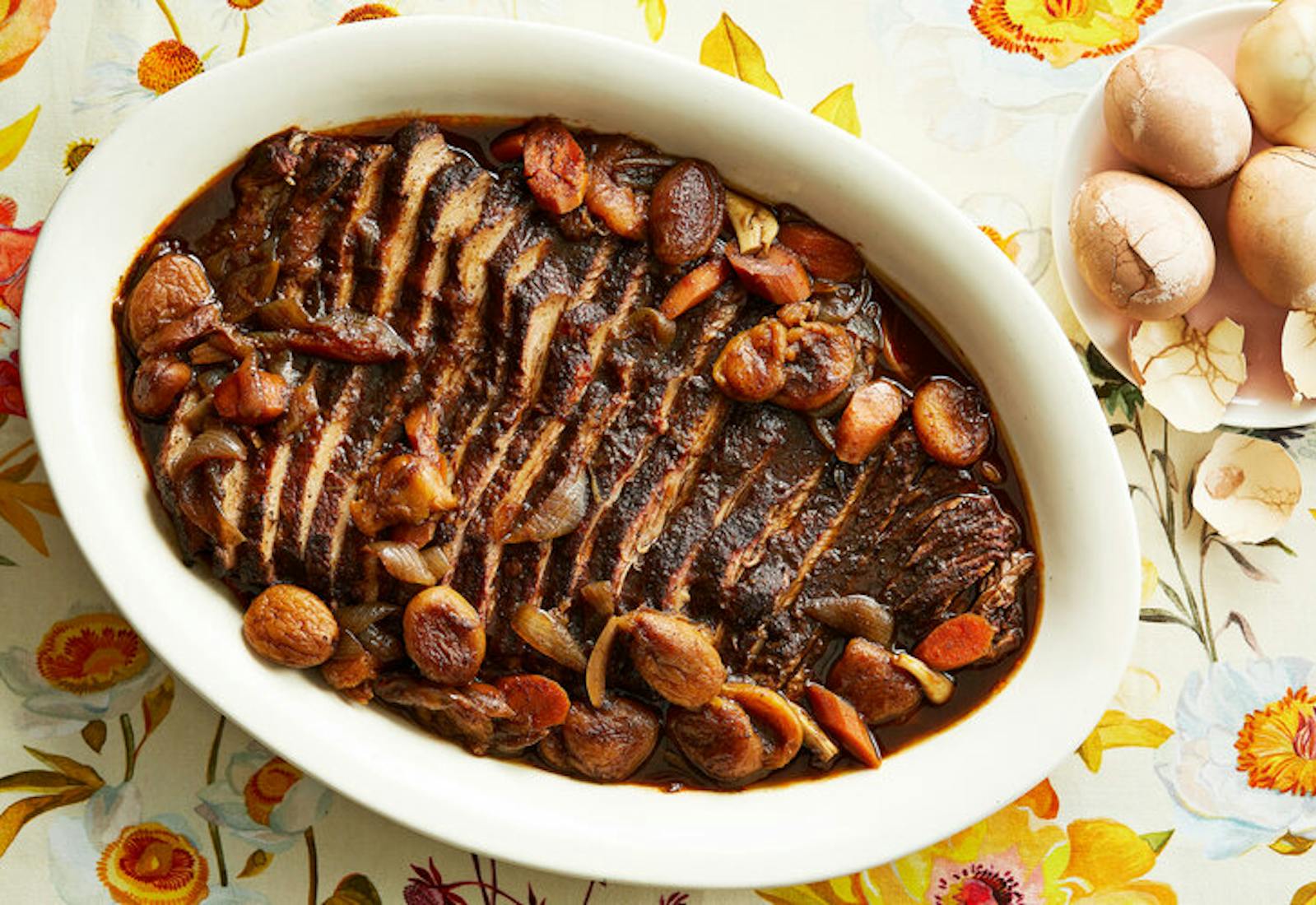 Sliced brisket in large casserole dish with whole eggs atop floral tablecloth.