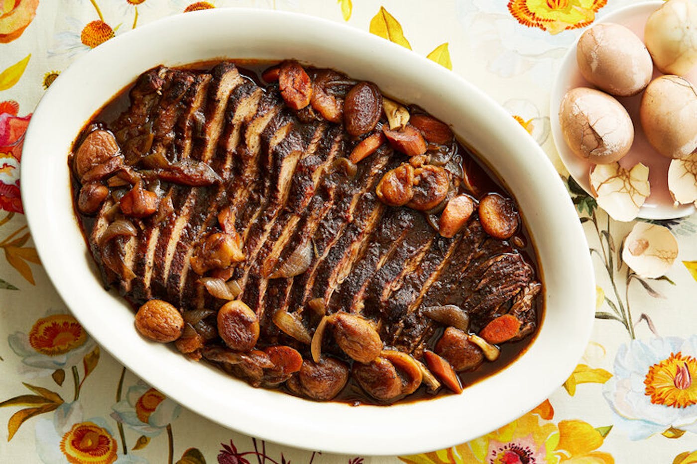 Sliced brisket in large casserole dish with whole eggs atop floral tablecloth.
