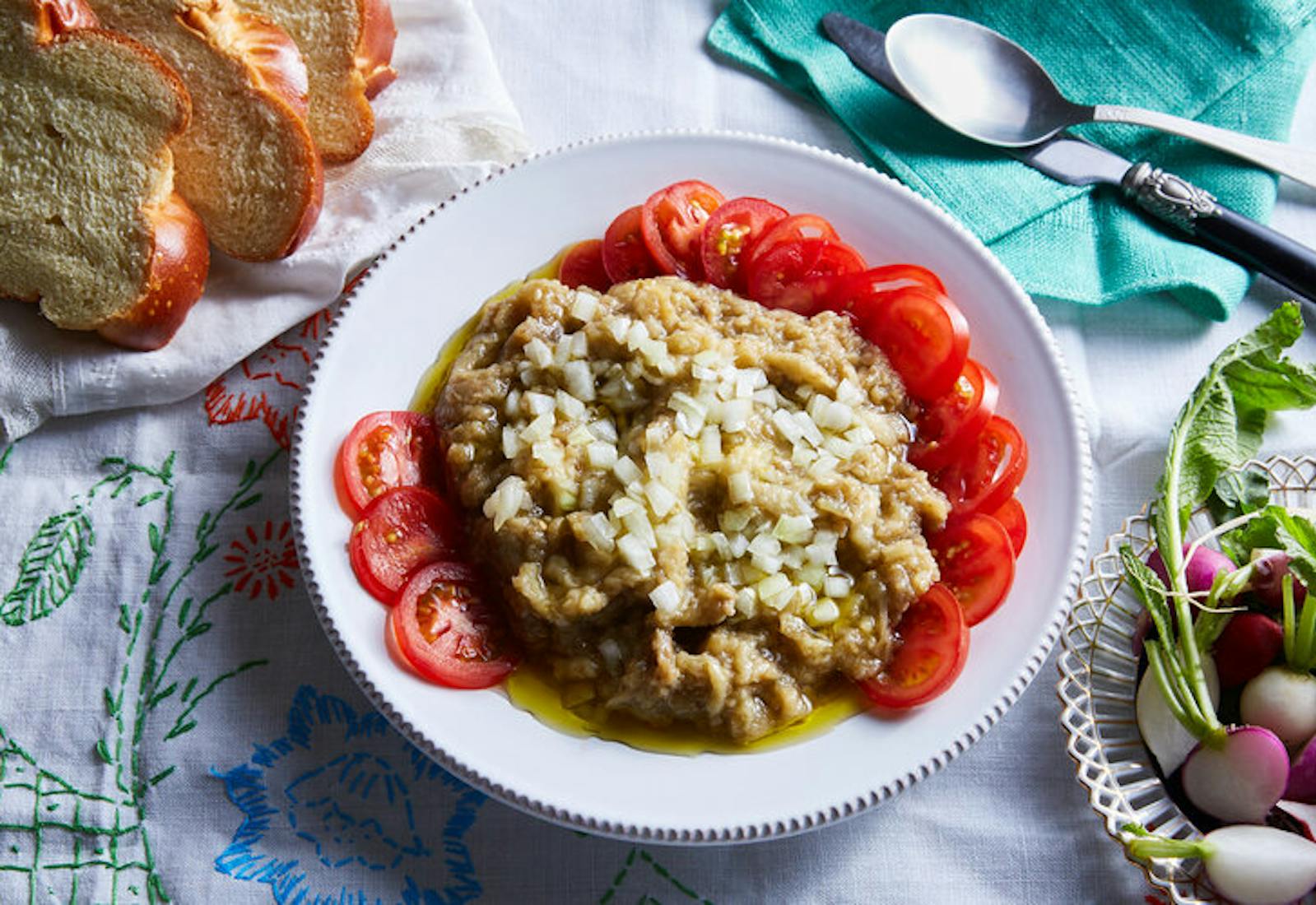 Eggplant salad with raw onions and tomatoes alongside sliced challah and bowl of halved radishes.