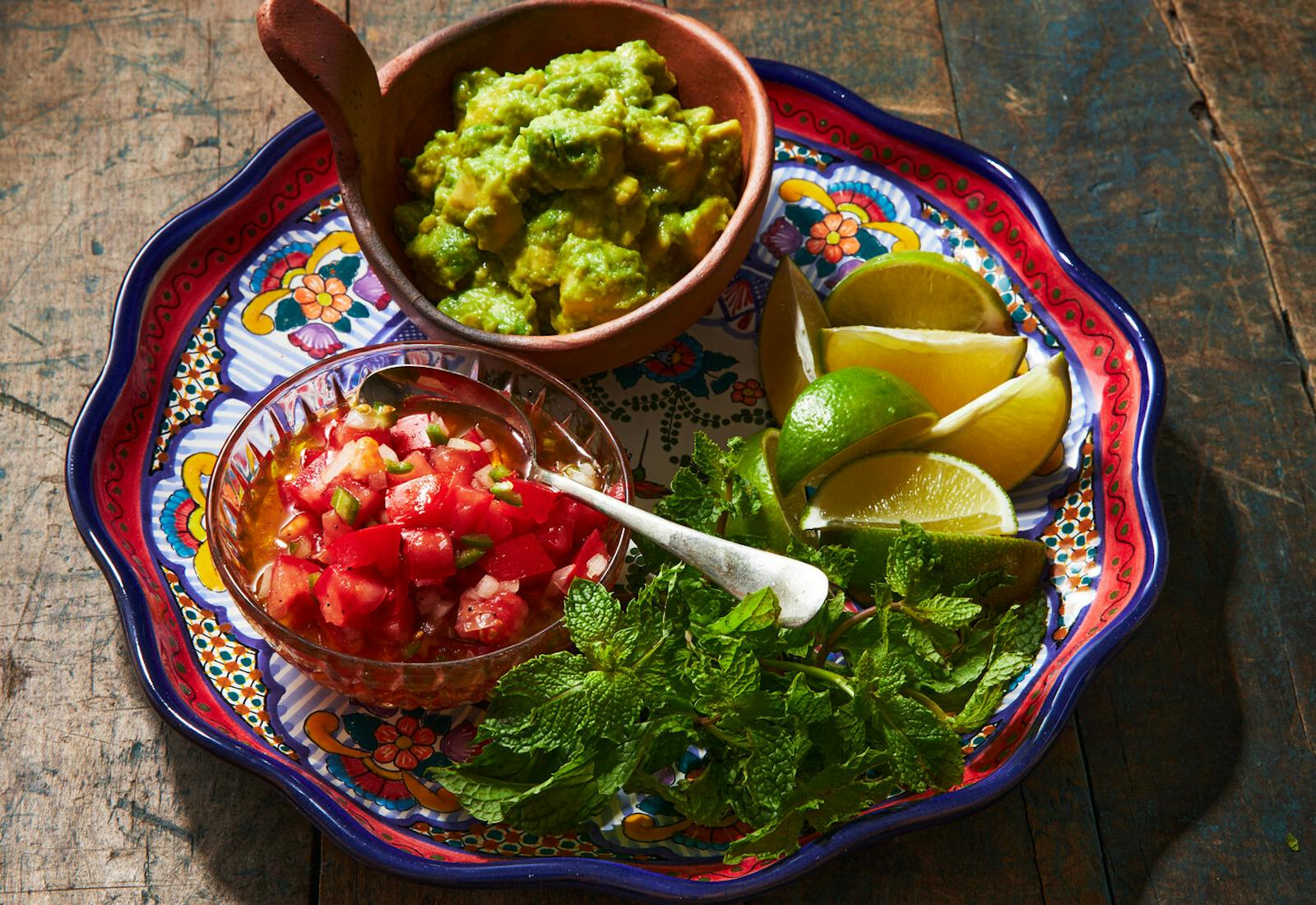 Salsa cruda in glass bowl alongside bowl of guacamole, lime wedges and mint on vibrant platter.