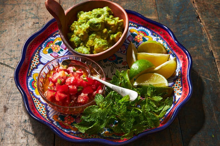 Salsa cruda in glass bowl alongside bowl of guacamole, lime wedges and mint on vibrant platter.