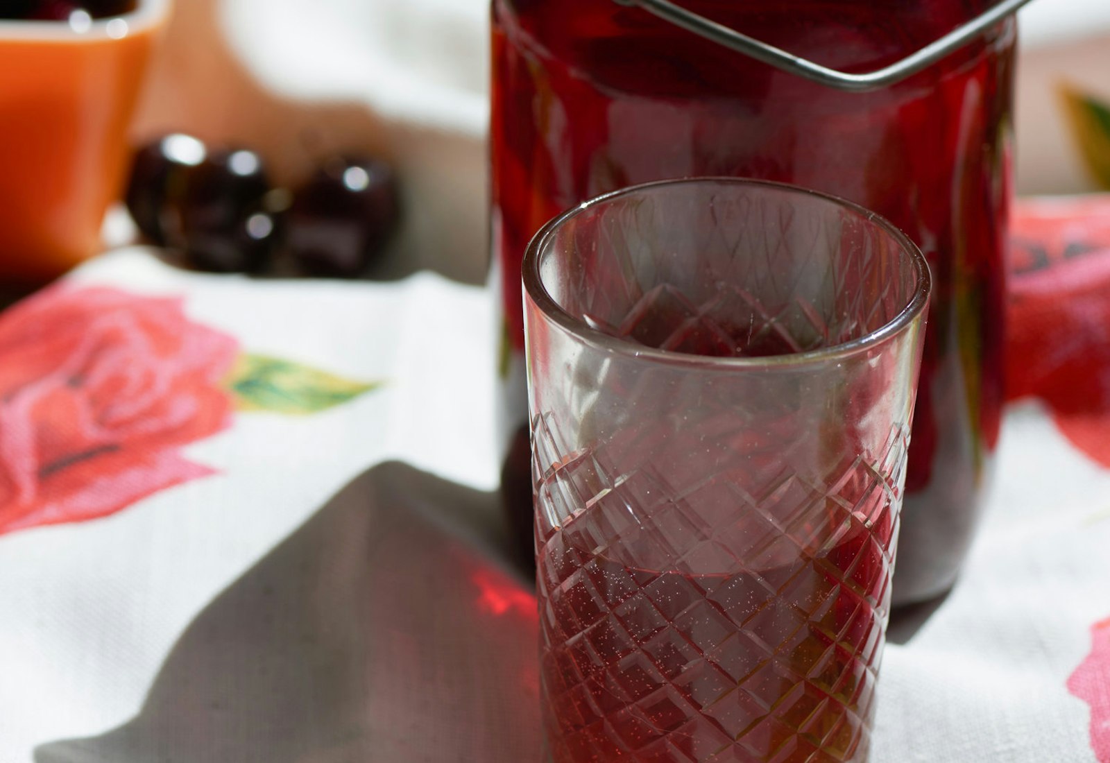 Kompot in jar and cup alongside dark red cherries atop floral tablecloth.