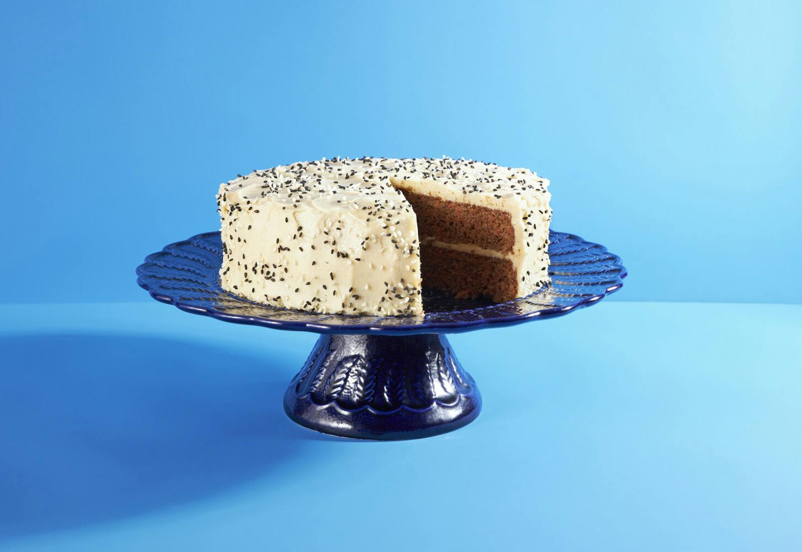 Tahini carrot cake with frosting and black and white sesame seeds on blue cake platter, blue background. 
