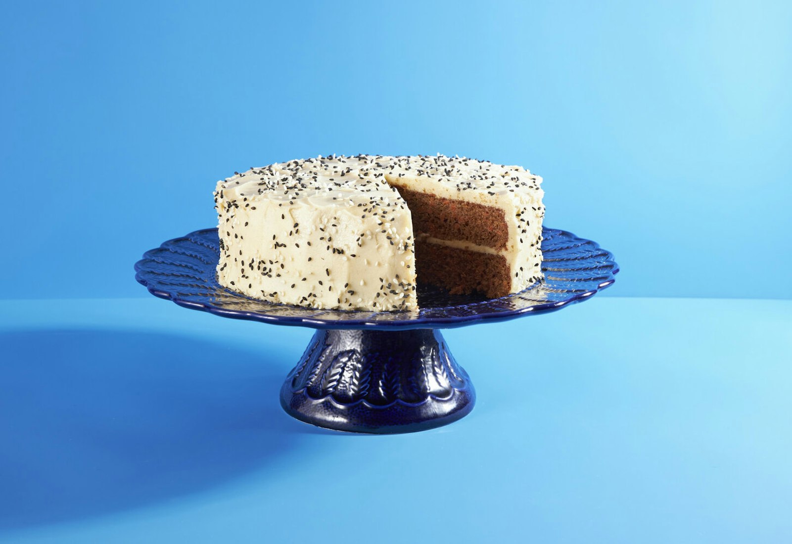 Tahini carrot cake with frosting and black and white sesame seeds on blue cake platter, blue background.