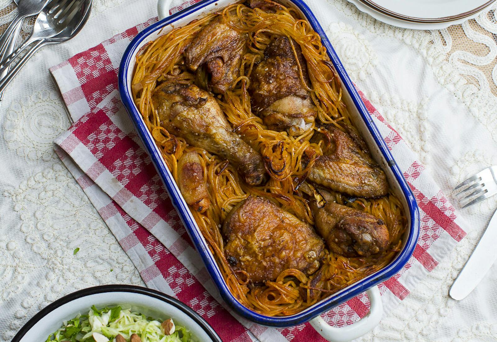 Macaroni chicken in blue casserole dish alongside cabbage salad with almonds and dish with lemon wedges.