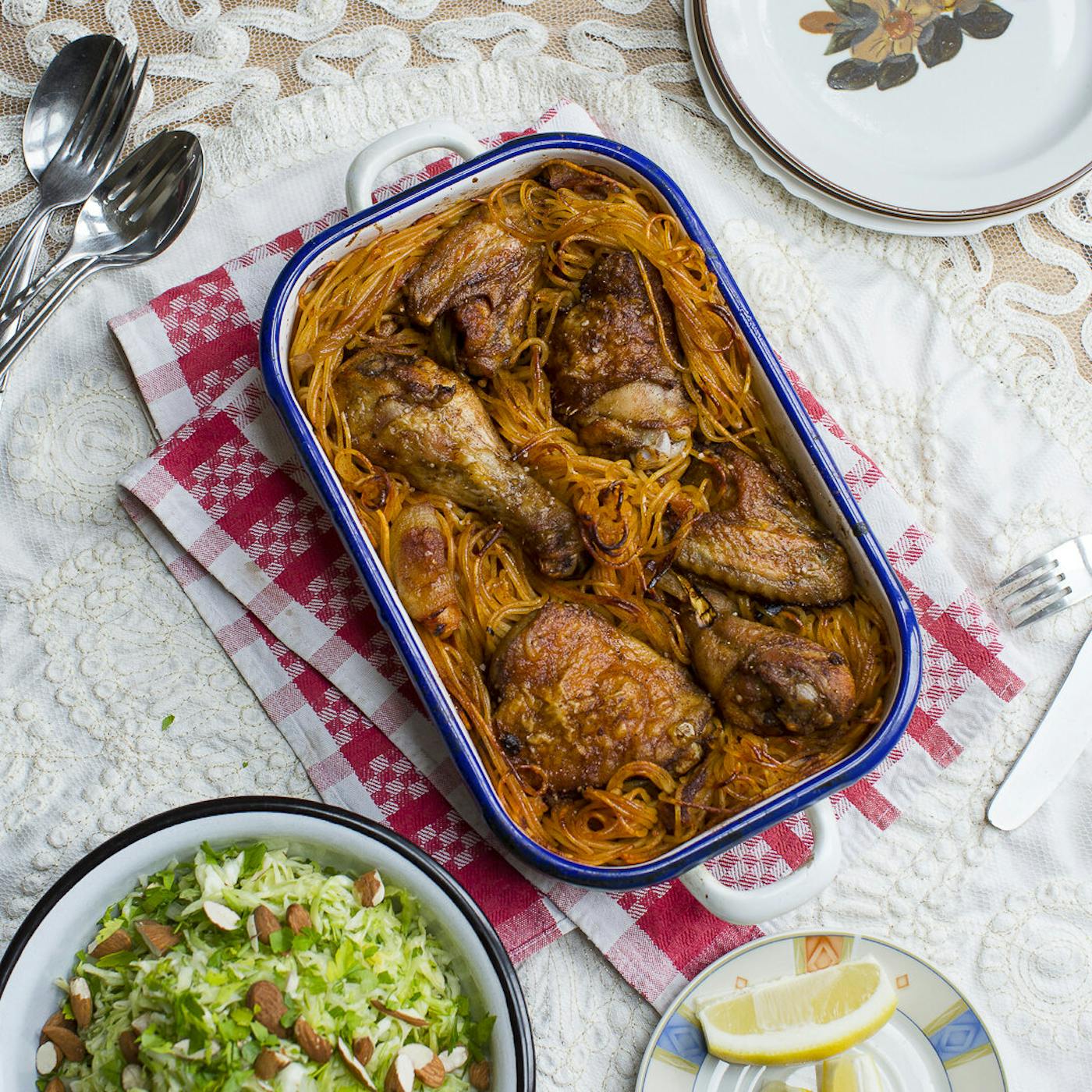 Macaroni chicken in blue casserole dish alongside cabbage salad with almonds and dish with lemon wedges.