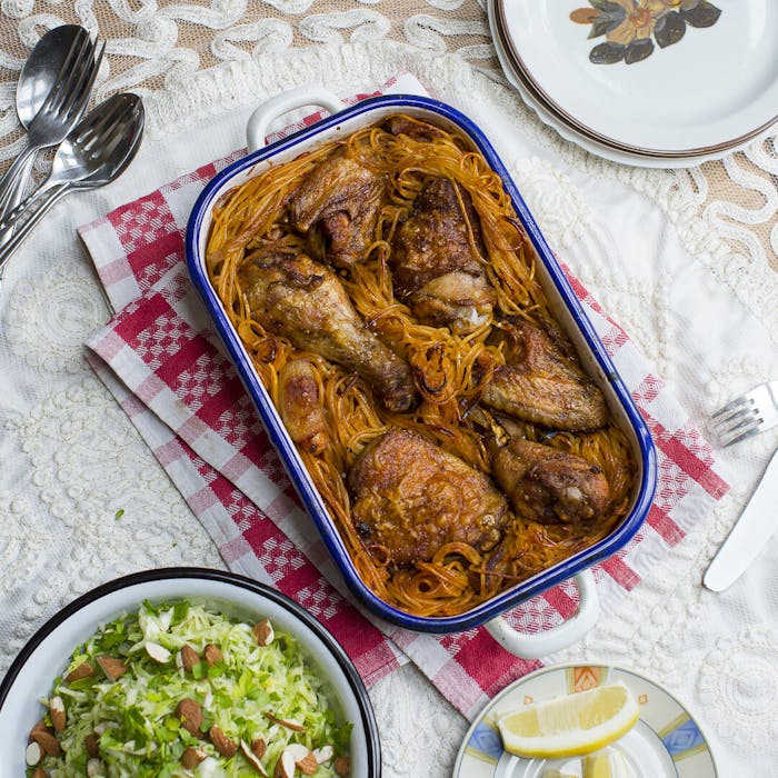 Macaroni chicken in blue casserole dish alongside cabbage salad with almonds and dish with lemon wedges.
