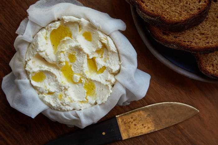 Cherkessian cheese straining over cheesecloth alongside plate of sliced bread atop wooden surface.