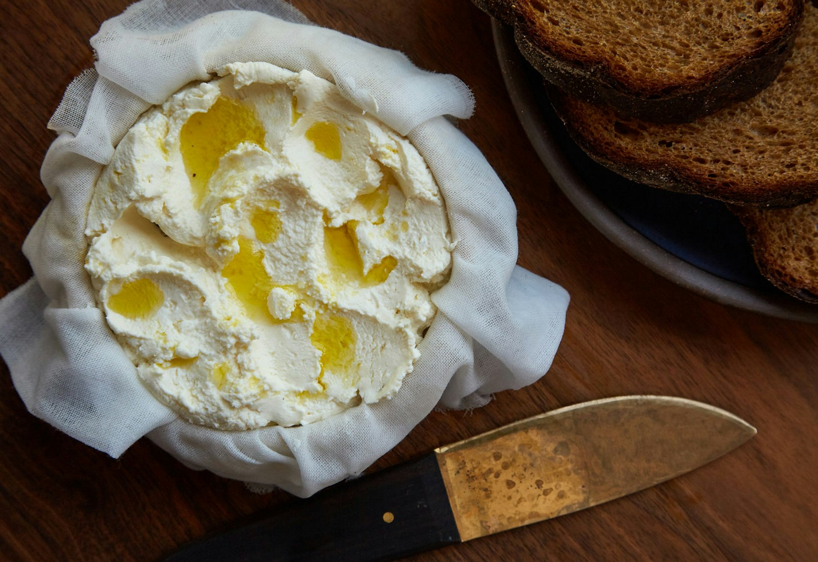 Cherkessian cheese straining over cheesecloth alongside plate of sliced bread atop wooden surface.