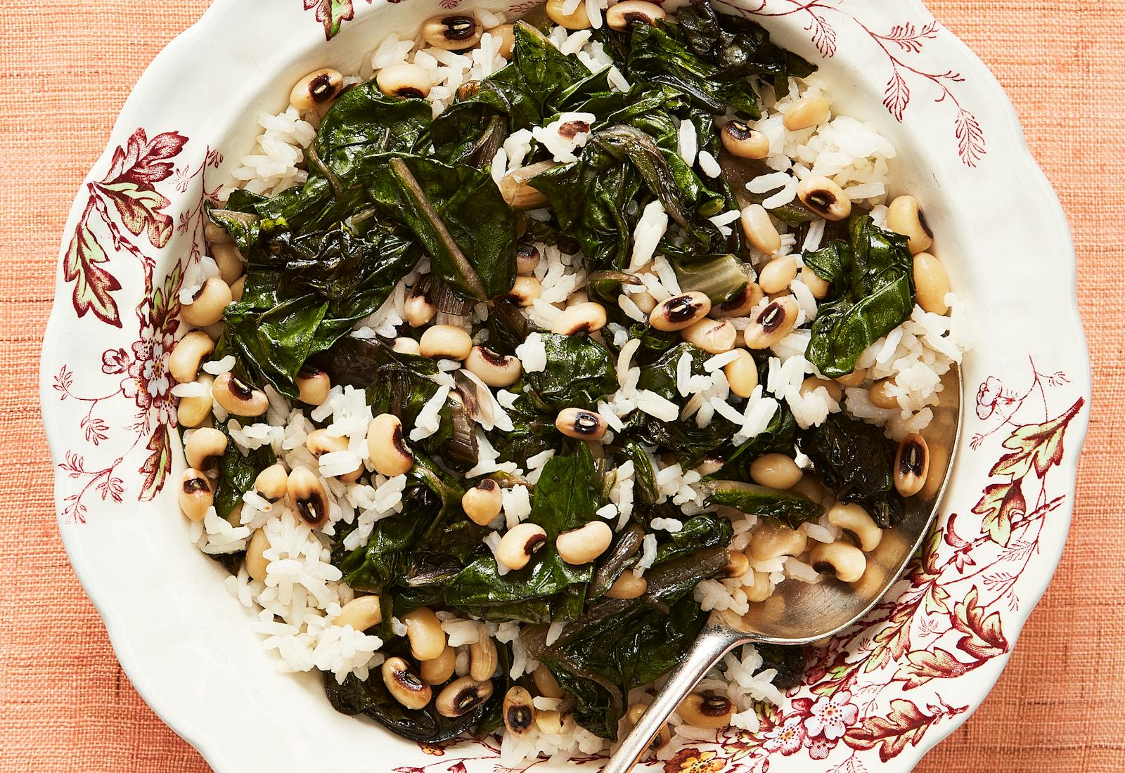 Rice with black eyed peas and chard in white floral bowl over salmon tablecloth.