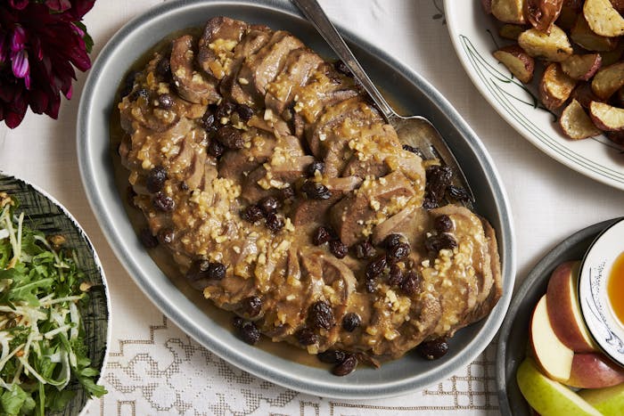 Sliced sweet-and-sour tongue with onions and raisins in large grey dish, alongside fresh flowers and apples with honey.