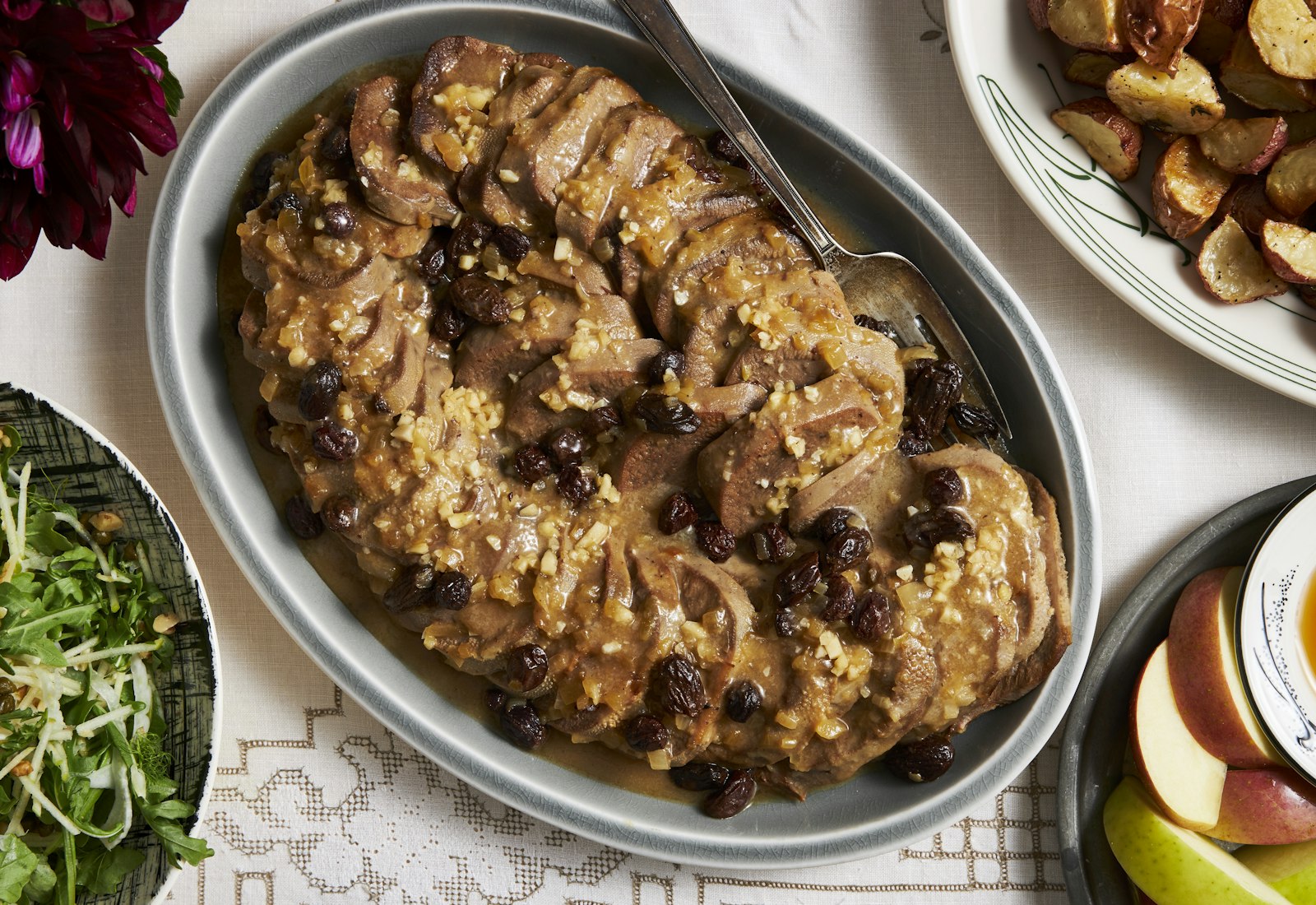 Sliced sweet-and-sour tongue with onions and raisins in large grey dish, alongside fresh flowers and apples with honey.