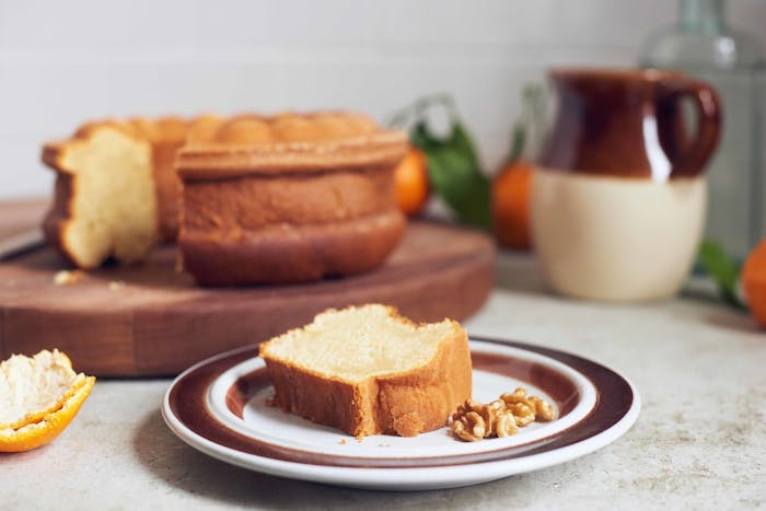 Slice of honey cake on dish with walnuts with entire cake on wooden cutting board, oranges and ceramic jug.