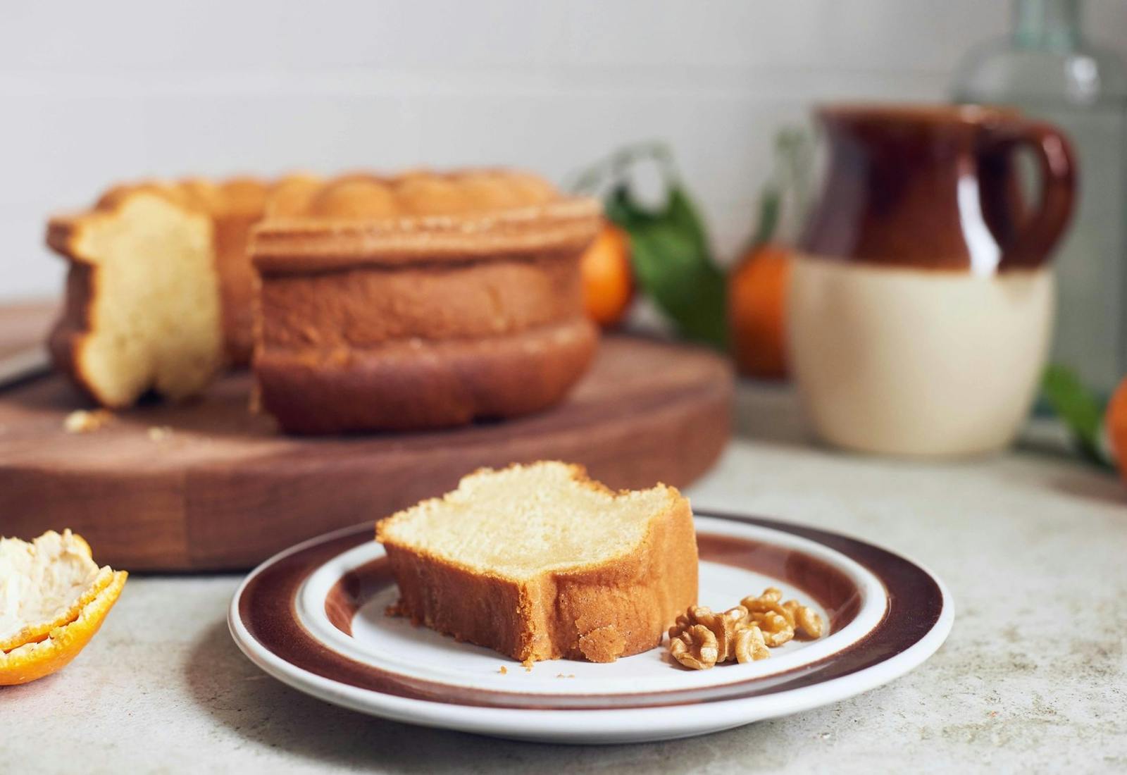 Slice of honey cake on dish with walnuts with entire cake on wooden cutting board, oranges and ceramic jug.