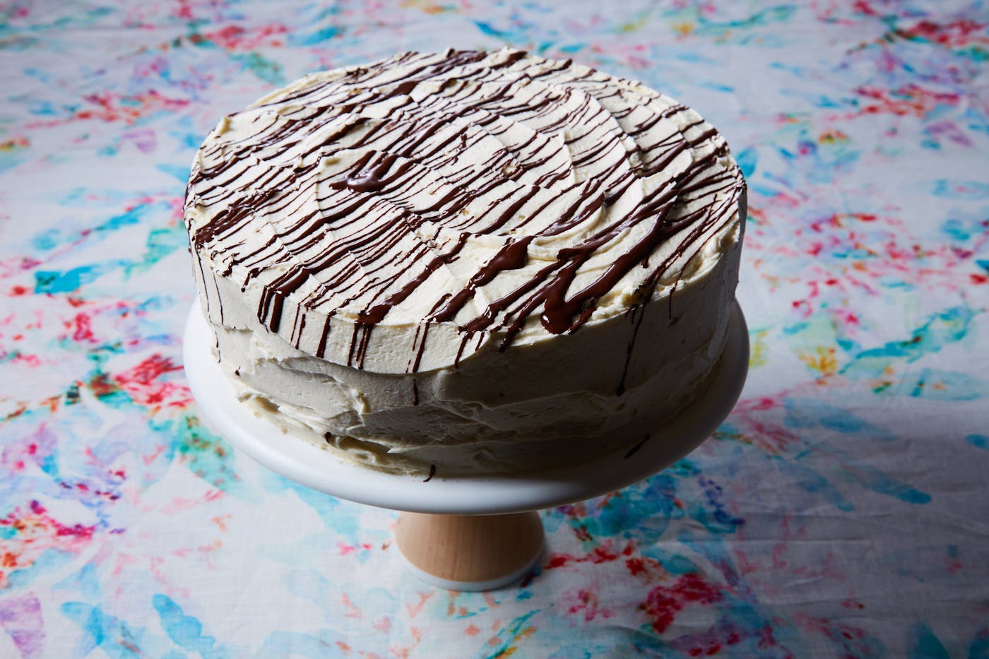 Chocolate cake with vanilla icing and chocolate drizzle on cake tray atop tie-dyed tablecloth.
