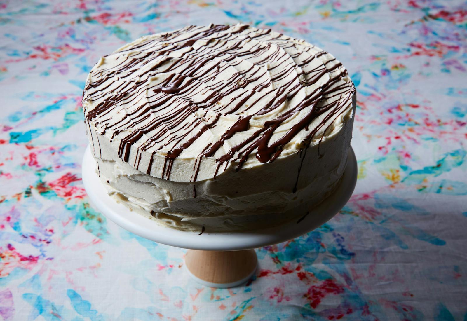 Chocolate cake with vanilla icing and chocolate drizzle on cake tray atop tie-dyed tablecloth.