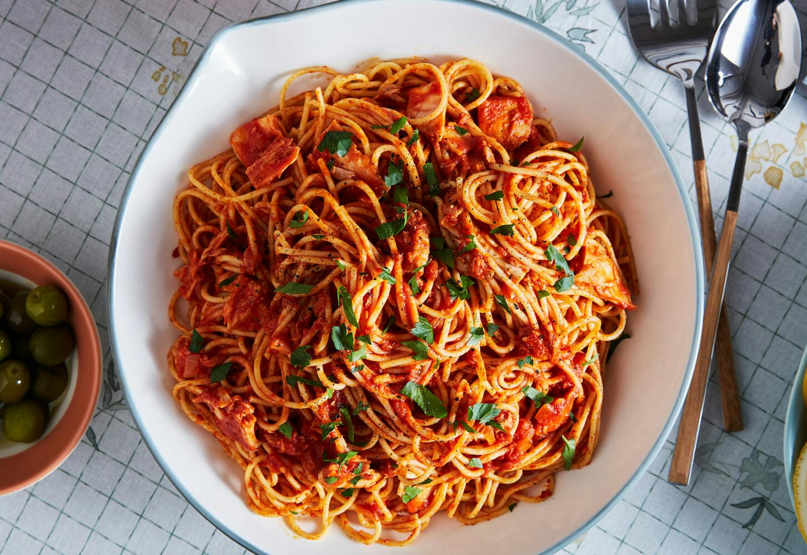 Spaghetti with tomatoes and tuna, topped with chopped parsley alongside bowls of olives and lemon wedges.