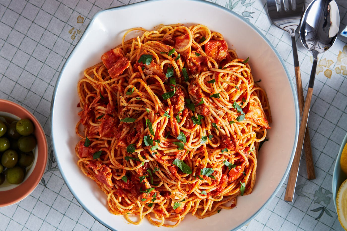 Spaghetti with tomatoes and tuna, topped with chopped parsley alongside bowls of olives and lemon wedges.
