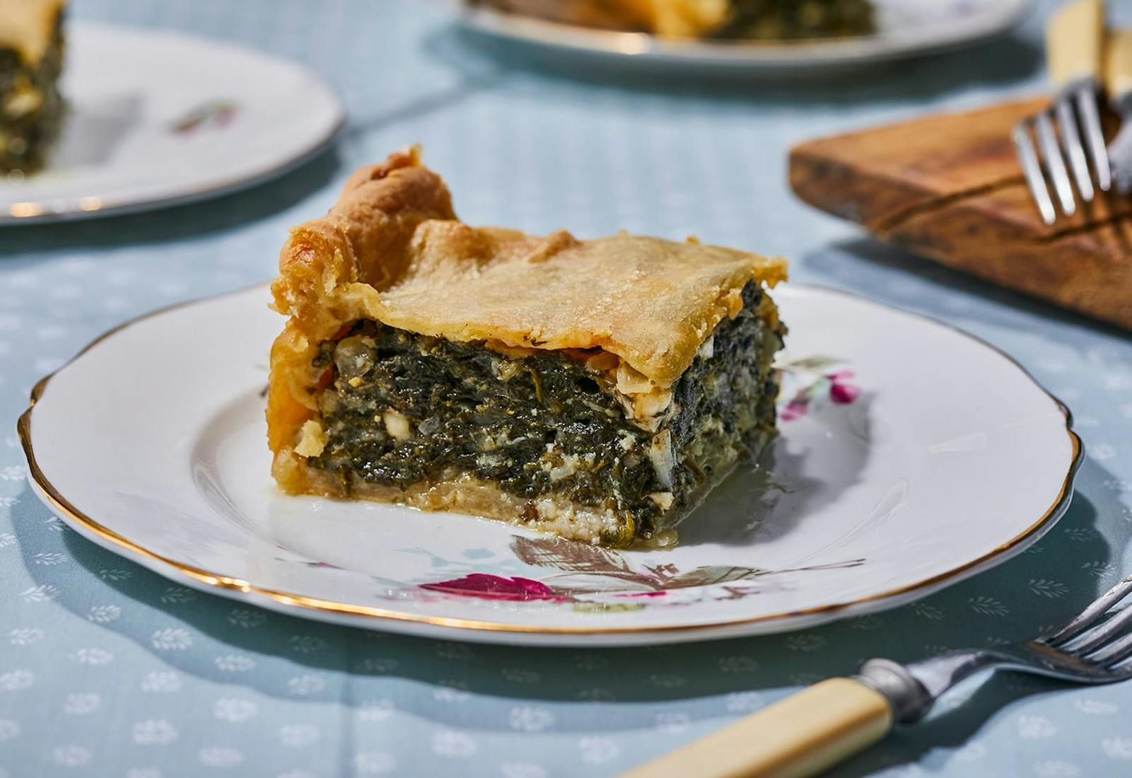 Slice of spanakopita on small dish, atop blue tablecloth.
