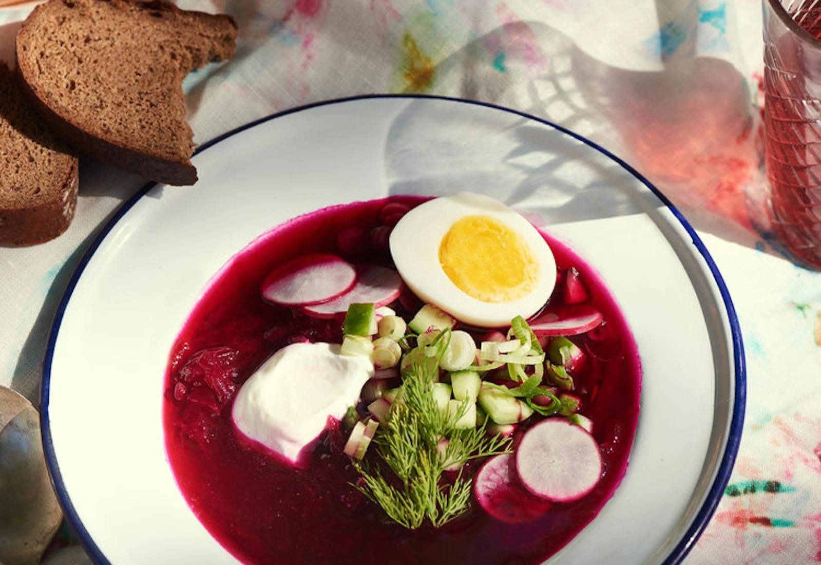 Bowl of svekolnik garnished with dill, radishes, cucumbers, scallions, sour cream and hard boiled egg alongside sliced brown bread atop tie-dyed tablecloth. 