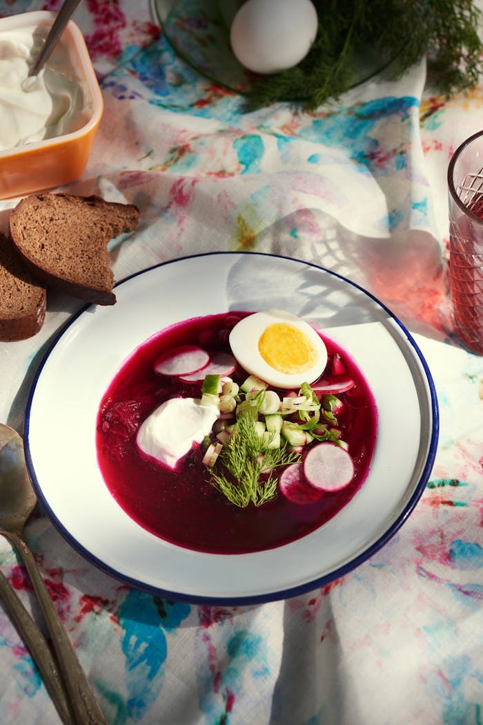 Bowl of svekolnik garnished with dill, radishes, cucumbers, scallions, sour cream and hard boiled egg alongside sliced brown bread atop tie-dyed tablecloth.