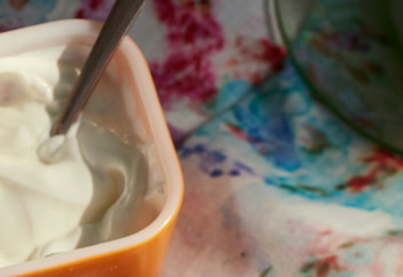 Bowl of svekolnik garnished with dill, radishes, cucumbers, scallions, sour cream and hard boiled egg alongside sliced brown bread atop tie-dyed tablecloth. 