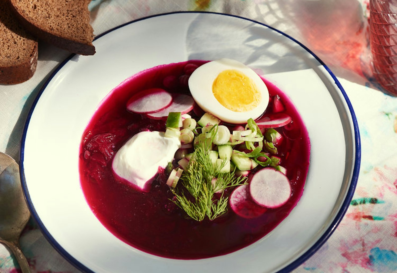 Bowl of svekolnik garnished with dill, radishes, cucumbers, scallions, sour cream and hard boiled egg alongside sliced brown bread atop tie-dyed tablecloth.