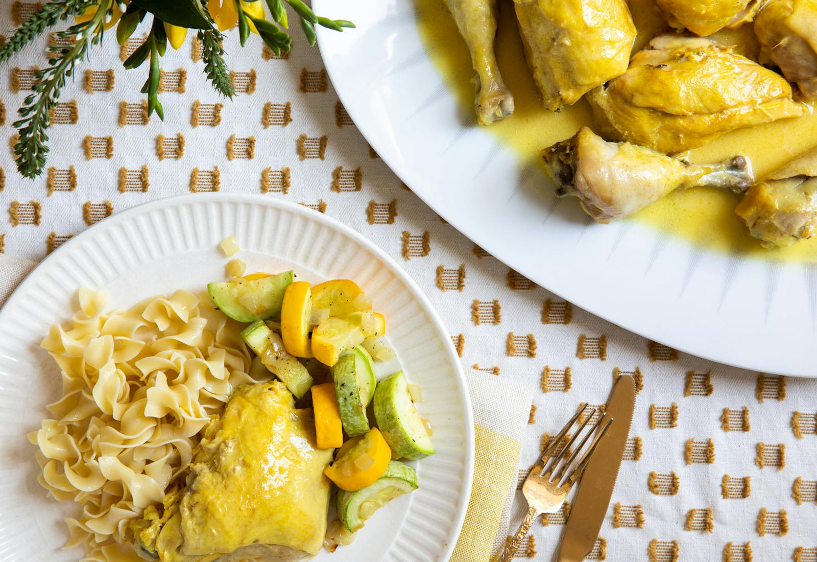 Chicken with lemon-saffron sauce with serving of chicken with roasted zucchini and pasta, atop white tablecloth with gold woven patterns.