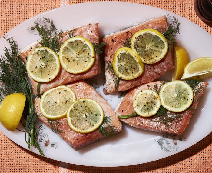 Poached salmon with sliced lemon and dill fronds on oval dish atop wicker surface.