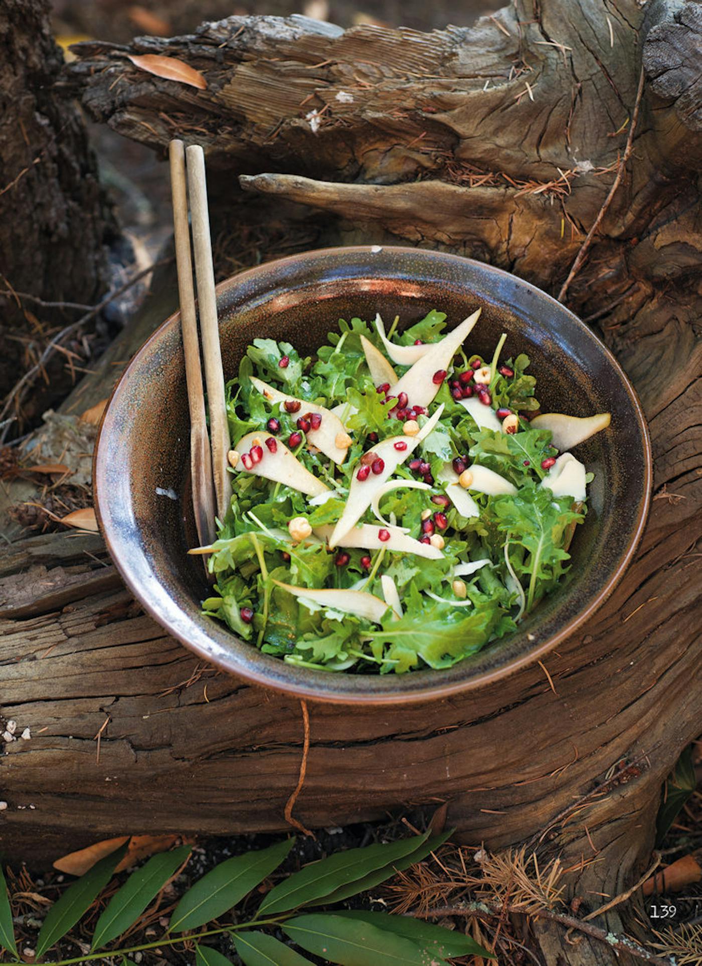 Kale hazelnut salad in bronze bowl with wooden serving utensils, outdoor setting atop tree roots.