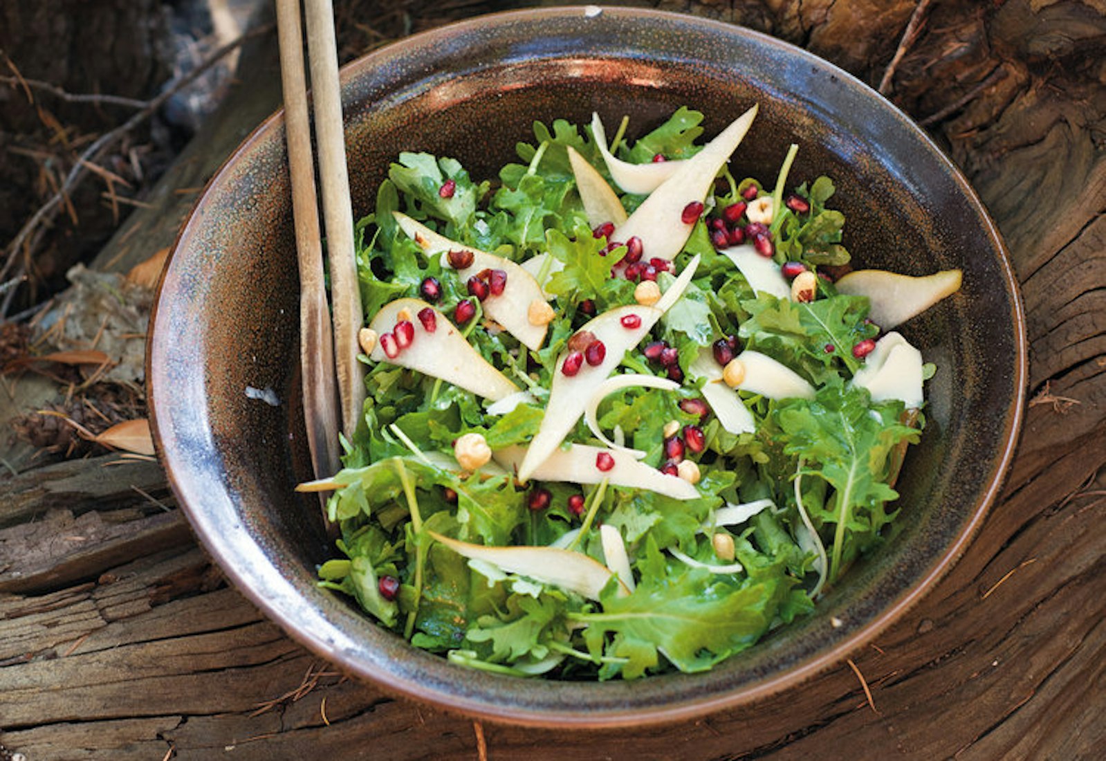 Kale hazelnut salad in bronze bowl with wooden serving utensils, outdoor setting atop tree roots.