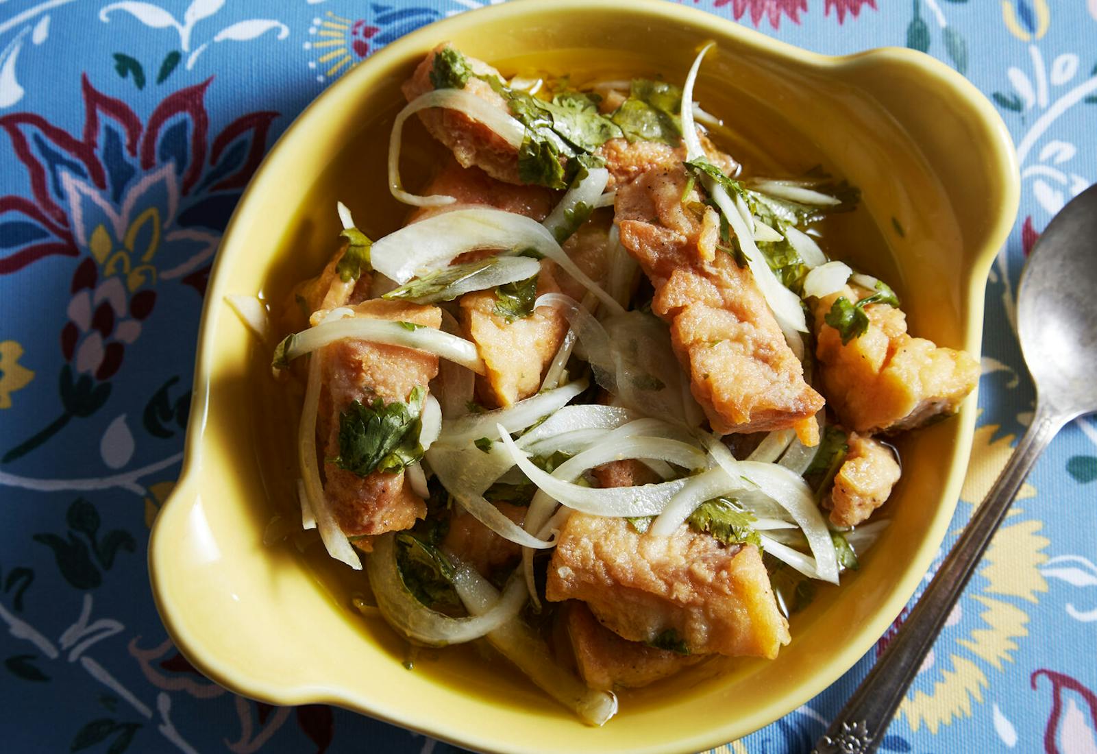 Escabeche with onion slices and cilantro in yellow bowl atop blue floral tablecloth.